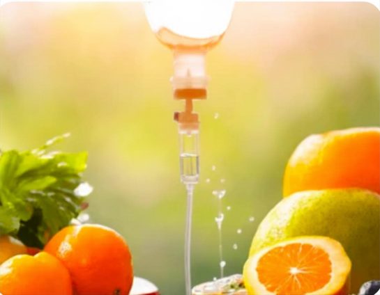 An infusion drip chamber in warm sunlight beside fresh oranges and greenery