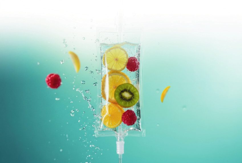 A clear IV bag holding slices of lemon, orange and kiwi with raspberries, suspended in bright turquoise water