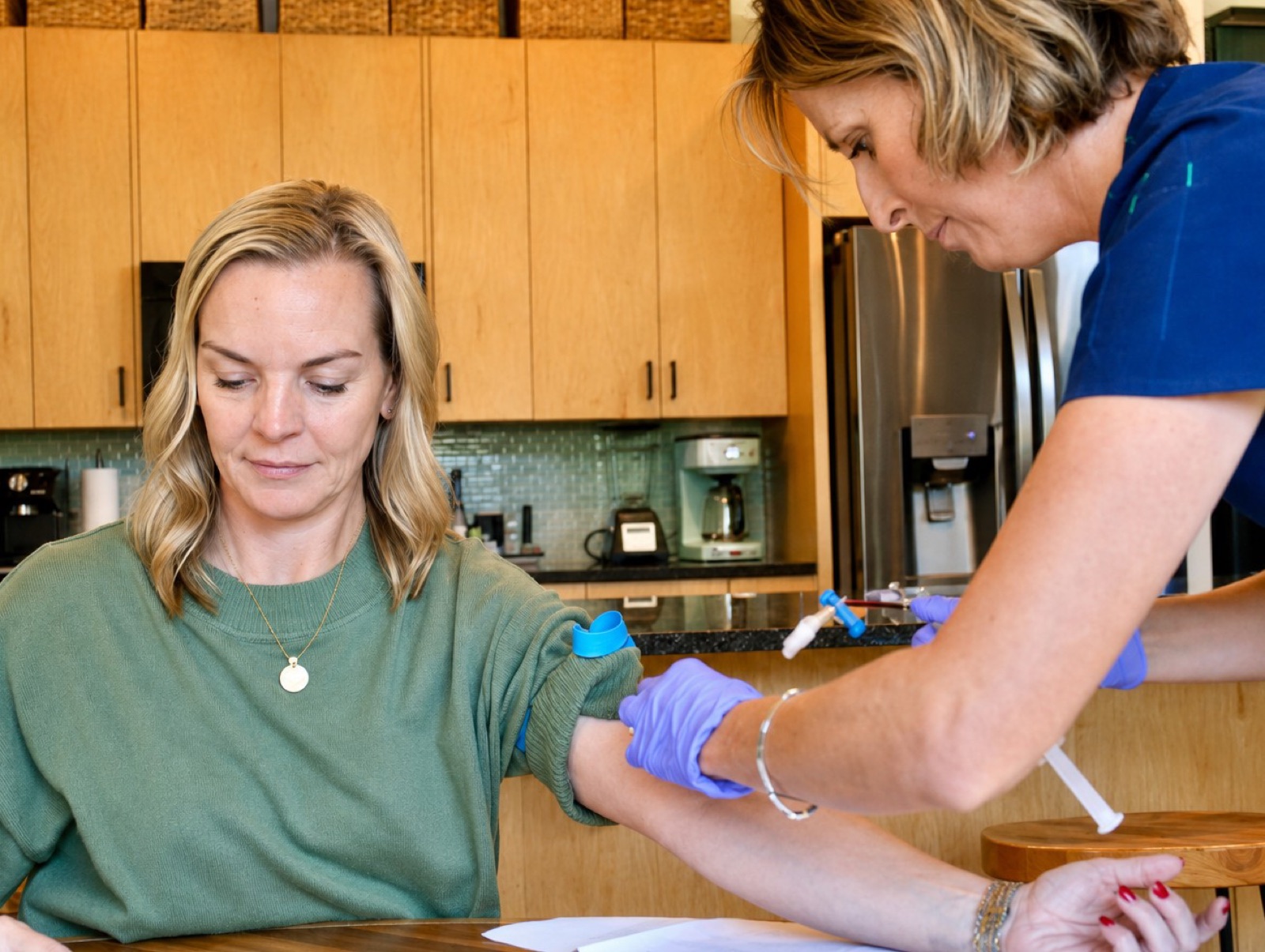A nurse in blue scrubs and gloves prepares an IV in a woman's arm at her kitchen table